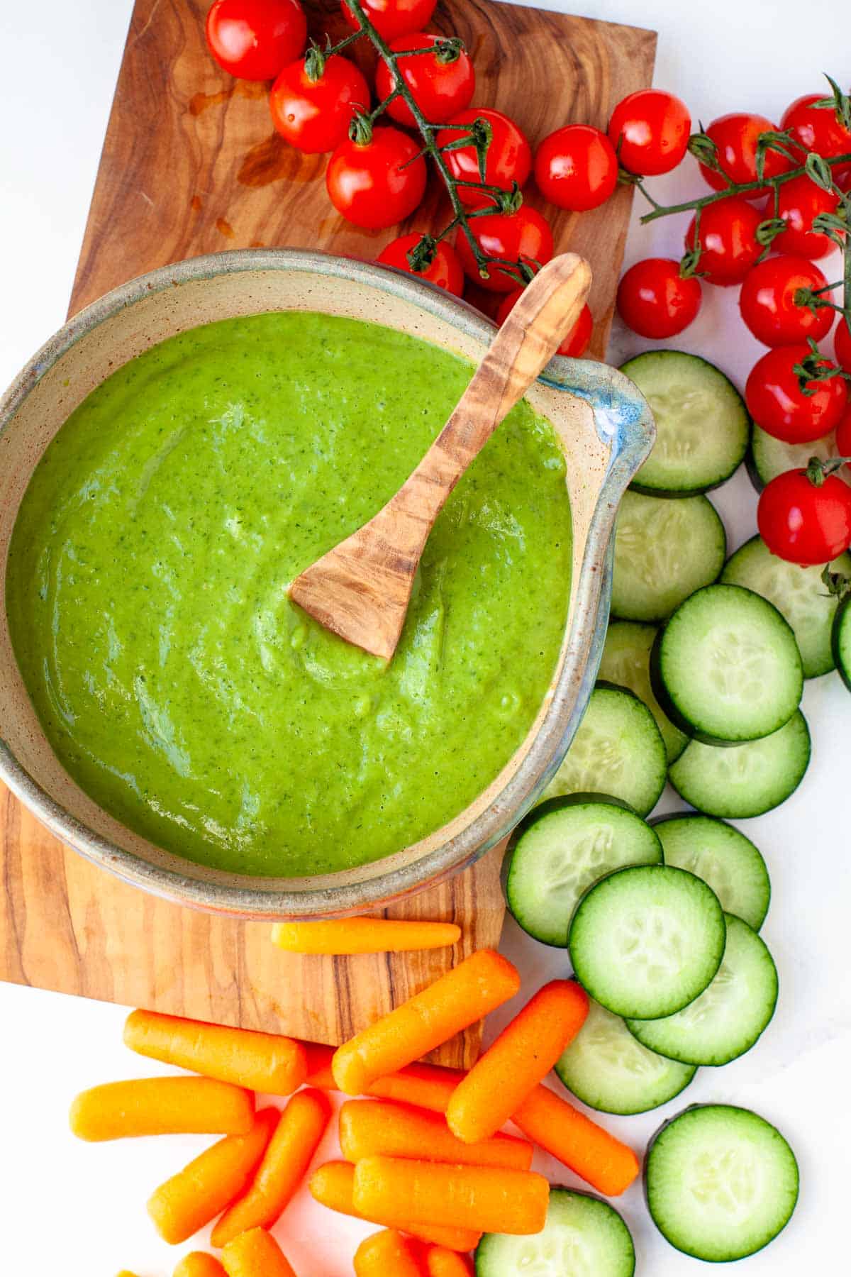 Vegan Green Goddess Dressing in serving bowl atop a wooden cutting board along with fresh cherry tomatoes, sliced cucumber, and baby carrots.