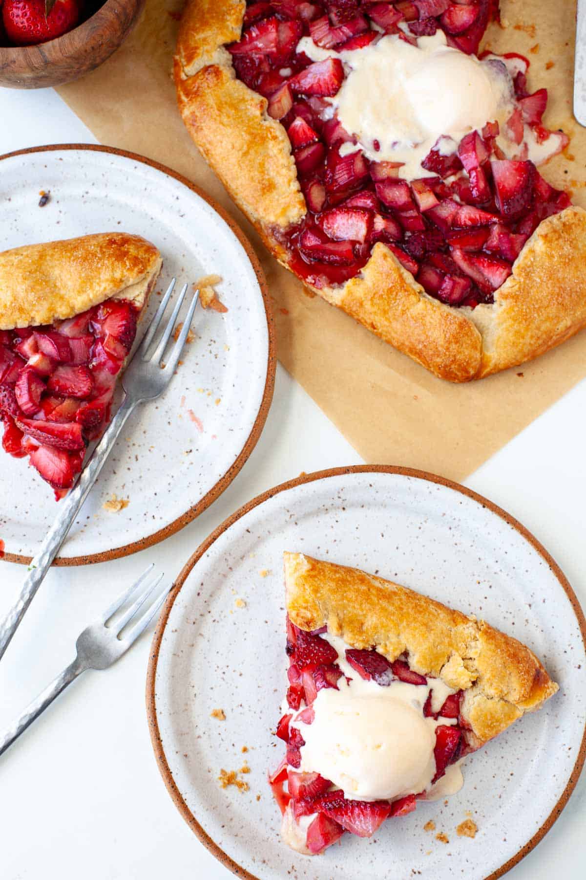 two white speckled plates with slices of strawberry rhubarb galette and whole galette with scoop of ice cream