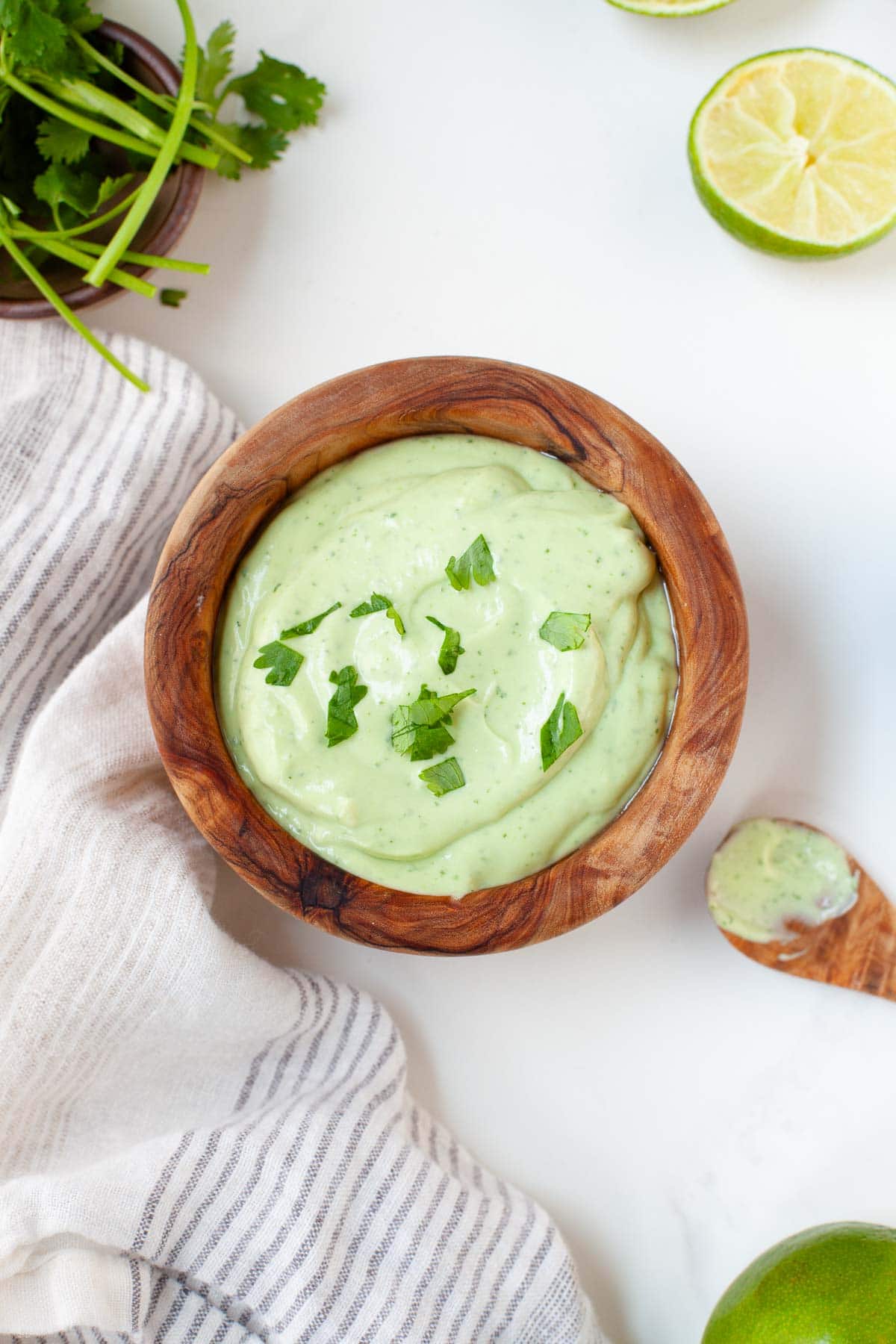 Close up of wooden bowl with avocado lime crema on white surface next to kitchen towel and wood spoon